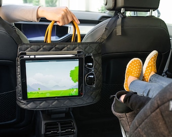 May include: Black quilted car seat organizer with a yellow handle, holding a tablet with a stylus, and sunglasses. The organizer is attached to the back of a car seat. A person's feet are resting on a car seat.