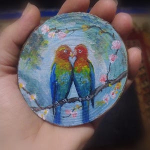 May include: A hand-painted, round wooden coaster featuring two colorful lovebirds perched on a branch with pink blossoms. The birds have red heads, yellow and green bodies, and blue wings, set against a blue background.