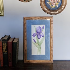 May include: A framed watercolor painting of a purple iris flower with green leaves and buds. The painting is set against a blue background and framed in a light brown wooden frame. Decorative plates hang on the wall above the painting.