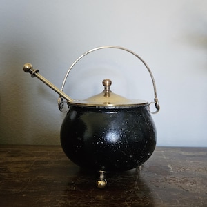 May include: A small, black, round pot with a silver handle and a gold-colored lid. A silver spoon rests on the side. The pot has three small gold-colored feet.
