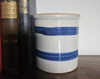May include: A cylindrical stoneware crock with a light gray base and two horizontal blue stripes. The rim and base are a light brown color. Two antique books are visible to the left of the crock.