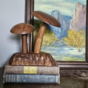 May include: A wooden mushroom sculpture with two mushroom caps and stems, mounted on a carved base. The sculpture sits atop two vintage books. A framed landscape painting hangs in the background.