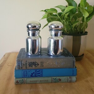 May include: Two silver metal salt and pepper shakers with rounded tops, sitting on a stack of books. The books are titled "Famous Seamen of America", "Imperial Woman", "Daniel Boone", "Pearls Buck", and "Webster Powers".