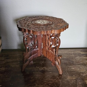 May include: A small, round, brown wooden folding stool featuring intricate floral carvings on the top surface and legs. The center of the tabletop displays a circular inlay with a white leaf pattern. The structure is supported by three carved legs.