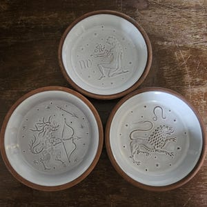 May include: Three white ceramic dishes with brown rims, each featuring a different zodiac sign: Virgo, Sagittarius, and Leo. The zodiac symbols are embossed in the center of each dish.