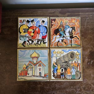 May include: Set of four decorative ceramic tiles with gold-colored frames. Each tile features a colorful, detailed illustration of Russian folk art scenes, including musicians, horsemen, a church, and a large cannon. The tiles are arranged on a dark wooden surface.