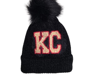 May include: Black knit beanie with a fluffy pom pom on top. The beanie has a gold and red "KC" logo with rhinestones.