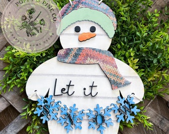 May include: White wooden snowman with a green hat, a colorful scarf, and a banner that reads "Let it snow". The snowman is decorated with blue snowflake cutouts.