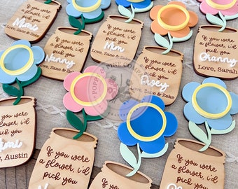 May include: A set of ten wooden flower mason jar ornaments with a variety of colors and names. Each ornament has a flower with a yellow center and a wooden mason jar with a message that says "If you were a flower, I'd pick you" followed by a name. The names include Mimi, Nanny, Mum, Dwey, Granny, Austin, Nana, Chachie.