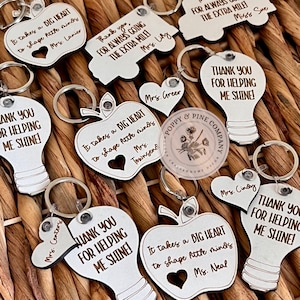 May include: A collection of wooden keychains with different designs, including an apple, a lightbulb, and a truck. Each keychain has a personalized message, such as "It takes a big heart to shape little minds" and "Thank you for helping me shine!" The keychains are made by Poppy & Pine Company.
