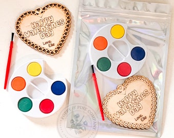 Puede incluir: Dos letreros de madera en forma de corazón para el Día de San Valentín con el texto "Happy Valentines Day" y "Love. Ellie". Cada letrero está empaquetado con una pequeña paleta de pintura y un pincel.