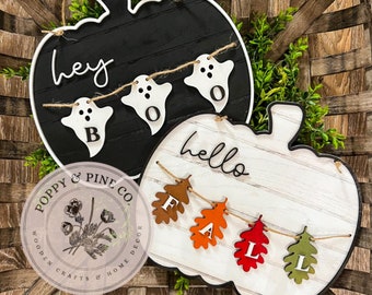 May include: Two wooden pumpkin-shaped signs with a black and white ghost garland on one and a brown, orange, and green leaf garland on the other. The signs say "hey boo" and "hello fall".