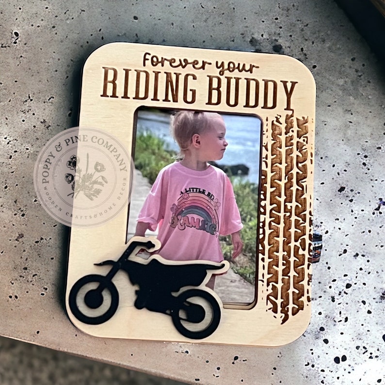 May include: A wooden photo frame with a cutout of a dirt bike and the text "Forever your Riding Buddy". The frame is light brown and has a photo of a young girl wearing a pink shirt with a rainbow graphic.