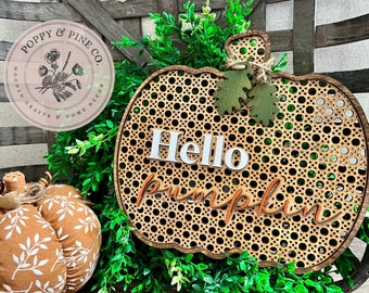 May include: A wooden pumpkin-shaped sign with a woven pattern and the words "Hello Pumpkin" in white and orange lettering. The sign is surrounded by green foliage.