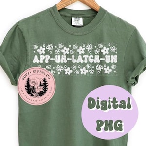Op de afbeelding: Een groen T-shirt met een witte afbeelding waarop "APP-UH-LATCH-UN" staat, met een bloemmotief boven en onder de tekst. Een roze cirkel met de tekst "POPPY & PINE CO. LASER ENGRAVED GIFTS & MORE" bevindt zich aan de linkerkant van het shirt.