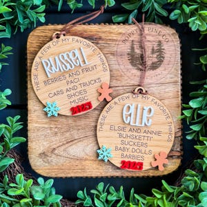 Puede incluir: Dos adornos navideños de madera con los nombres "Russell" y "Ellie" en blanco, junto con cosas favoritas y el año 2025. Cada adorno tiene un copo de nieve y un acento de hombre de jengibre, y una cuerda marrón para colgar.