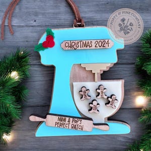 Könnte beinhalten: Ein blauer und hölzerner Weihnachtsanhänger in Form einer Küchenmaschine mit einem Nudelholz und dem Text "Mimi & Pop's Perfect Batch" und "Christmas 2024". Die Schüssel der Küchenmaschine enthält vier Lebkuchenmänner mit den Namen "Sophia", "Scarlett", "Ellie" und "Russell".