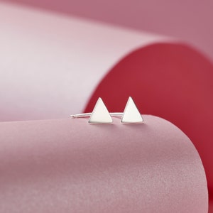 May include: A pair of silver triangle stud earrings on a pink background.