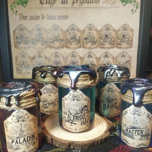 Op de afbeelding: Zes kleine glazen flesjes met zwarte was en touw rond de bovenkant. Elk flesje heeft een ander etiket met de naam van een fantasy-personage: Paladin, Clerigo, Mago, Master en twee anderen. De flesjes staan op een houten onderzetter.