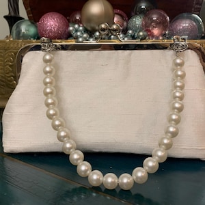 Puede incluir: Bolso de mano de satén marfil con asa de perlas. El cierre es plateado. El bolso es rectangular y tiene un diseño sencillo y elegante. El asa de perlas está hecha de perlas grandes, redondas y blanquecinas. El fondo incluye adornos decorativos.