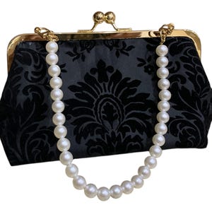 Może przedstawiać: Czarna aksamitna kopertówka ze złotym zapięciem i paskiem z pereł. Kopertówka ma kwiatowy wzór damasceński. Zapięcie i mocowania łańcuszka są w kolorze złotym. Pasek z pereł składa się z białych, okrągłych koralików.