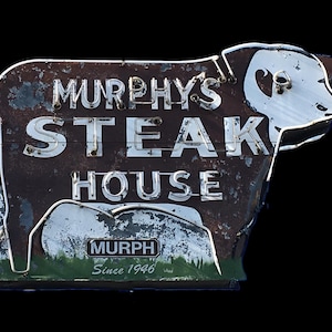 Op de afbeelding: Een vintage neon-bord in de vorm van een koe met de tekst "Murphy's Steak House" en "Since 1946" in witte letters op een bruine achtergrond.