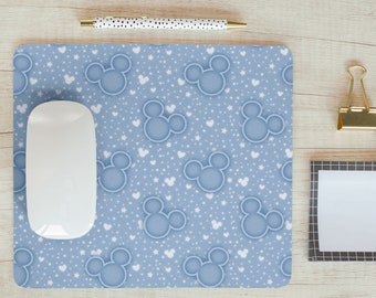May include: A blue mouse pad with a white Mickey Mouse head pattern. The mouse pad is on a light wood surface with a white computer mouse, a pen, and a gold paper clip.
