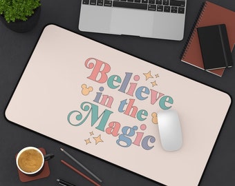 May include: A pink and white desk mat with the text "Believe in the Magic" in colorful retro font. The text is surrounded by stars and a Mickey Mouse icon. A white computer mouse is on the mat.