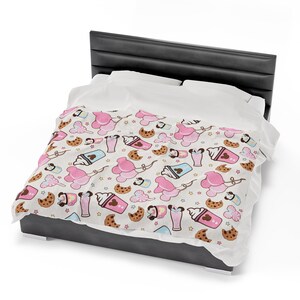 Op de afbeelding: Een wit bedovertrek met een patroon van roze Mickey Mouse oren, bruine koekjes en roze en bruine milkshakes. Het bedovertrek ligt op een grijs bedframe.
