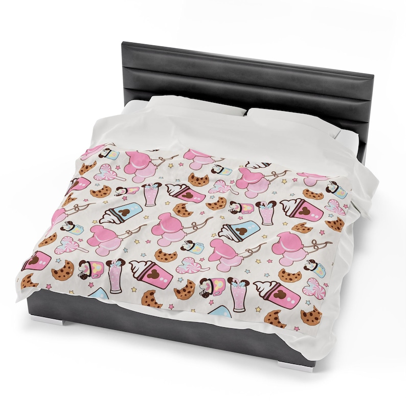 Op de afbeelding: Een wit bedovertrek met een patroon van roze Mickey Mouse oren, bruine koekjes en roze en bruine milkshakes. Het bedovertrek ligt op een grijs bedframe.