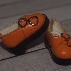 May include: A pair of orange leather doll shoes with heart-shaped buckles. The shoes have black soles and are made for a small doll.