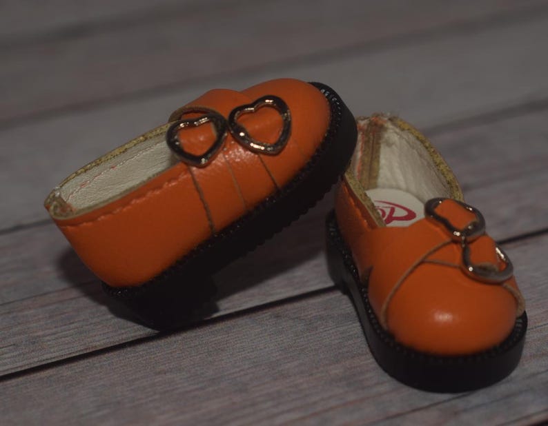 May include: A pair of orange leather doll shoes with heart-shaped buckles. The shoes have black soles and are made for a small doll.