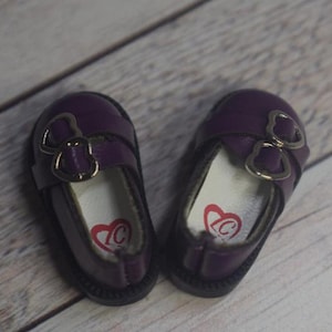 May include: A pair of purple doll shoes with silver heart-shaped buckles. The shoes have a white lining with a red heart logo.