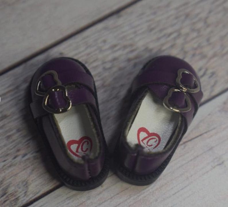 May include: A pair of purple doll shoes with silver heart-shaped buckles. The shoes have a white lining with a red heart logo.