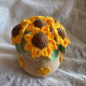 May include: A crocheted sunflower arrangement in a beige pot. The sunflowers have yellow petals and brown centres, with green leaves. The pot is decorated with yellow hearts.