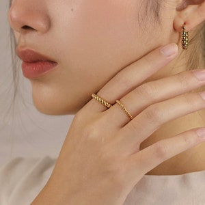 May include: Two gold rings on a person's finger. The ring on the index finger is a beaded ring. The ring on the middle finger is a thin, twisted ring.