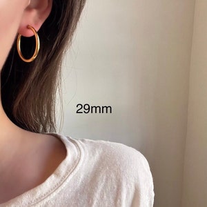 以下が含まれることがあります： 直径29mmのゴールドフープピアス。ピアスは人の耳につけられています。