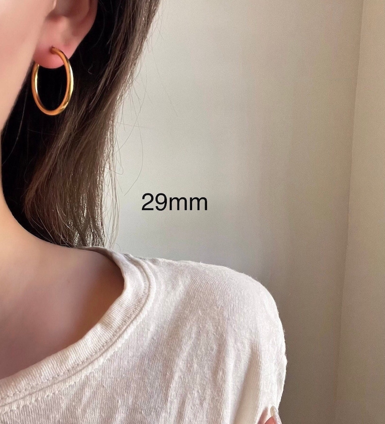 以下が含まれることがあります： 直径29mmのゴールドフープピアス。ピアスは人の耳につけられています。
