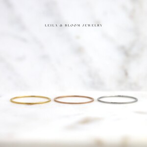 Puede incluir: Tres anillos finos apilables de oro, oro rosa y plata sobre un fondo de m&aacute;rmol blanco.  LEILA & BLOOM JEWELRY est&aacute; escrito en negro sobre los anillos.