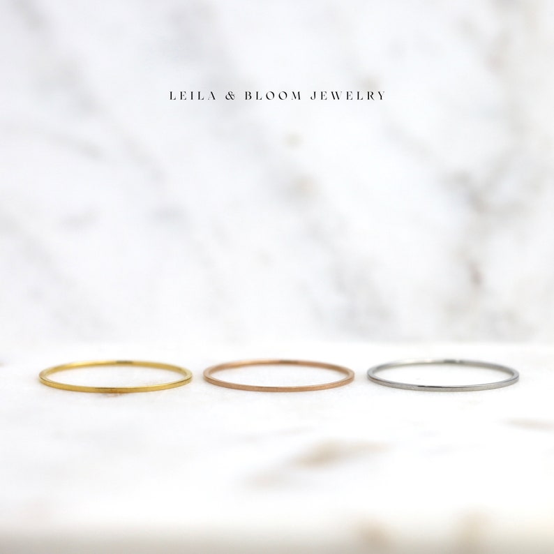 Puede incluir: Tres anillos finos apilables de oro, oro rosa y plata sobre un fondo de m&aacute;rmol blanco.  LEILA & BLOOM JEWELRY est&aacute; escrito en negro sobre los anillos.