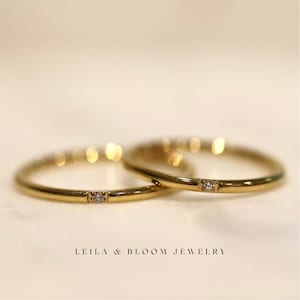 Puede incluir: Dos alianzas finas de oro con un peque&ntilde;o diamante engastado en cada anillo. Las alianzas est&aacute;n superpuestas.  LEILA & BLOOM JEWELRY est&aacute; escrito debajo de las alianzas.