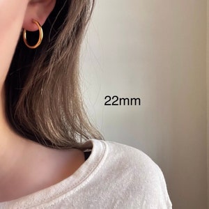 以下が含まれることがあります： 直径22mmのゴールドフープピアス。ピアスは人の耳につけられています。