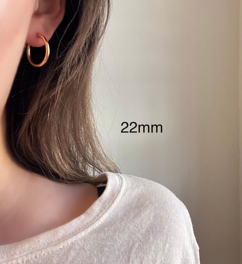 以下が含まれることがあります： 直径22mmのゴールドフープピアス。ピアスは人の耳につけられています。