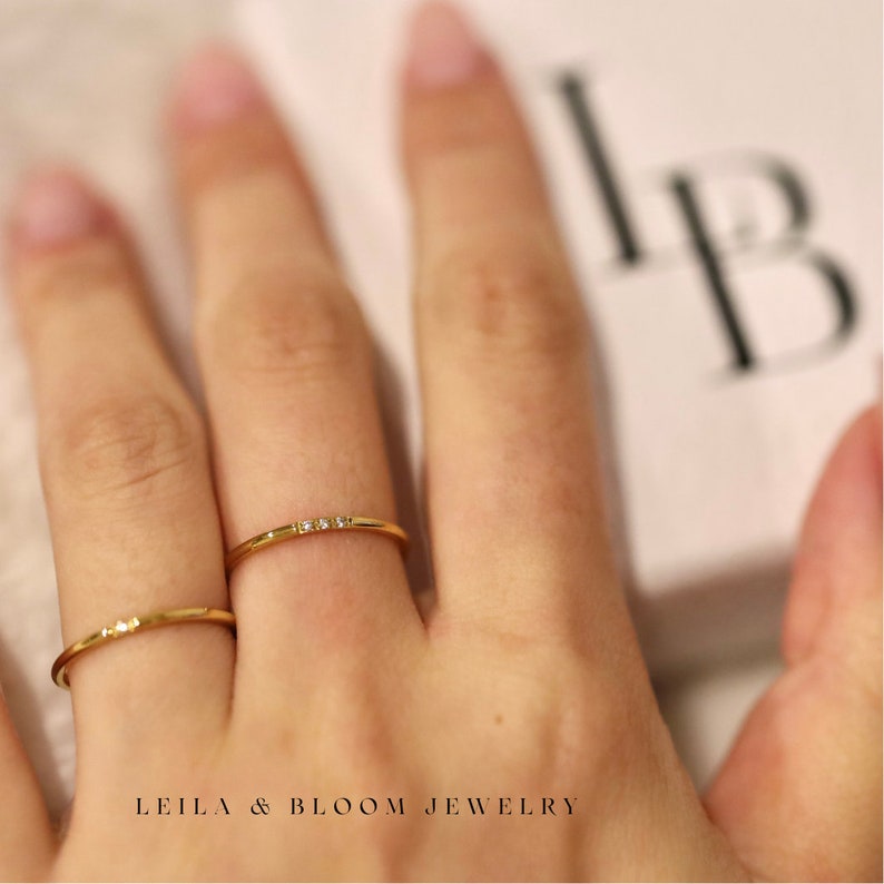 Puede incluir: Dos anillos delicados de oro con peque&ntilde;os diamantes engastados en fila. Los anillos se llevan en el &iacute;ndice y el dedo medio de una mano.