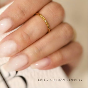 Puede incluir: Un delicado anillo de oro con un peque&ntilde;o diamante engastado en la banda. El anillo se lleva en el dedo de una mano con esmalte de u&ntilde;as rosa claro.