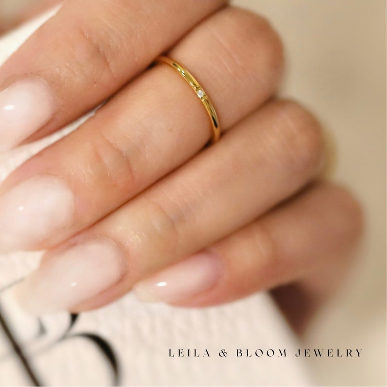 Puede incluir: Un delicado anillo de oro con un peque&ntilde;o diamante engastado en la banda. El anillo se lleva en el dedo de una mano con esmalte de u&ntilde;as rosa claro.