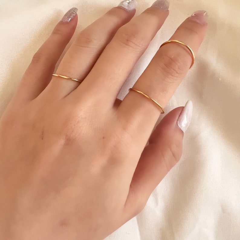 Puede incluir: Una mano con cuatro anillos finos de oro. Los anillos son simples y delicados, con un acabado liso.