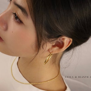 以下が含まれることがあります： 女性が金のチェーンネックレスと金のフープピアスをしています。画像の右下隅に「LEILA & BLOOM JEWELRY」というテキストが表示されています。