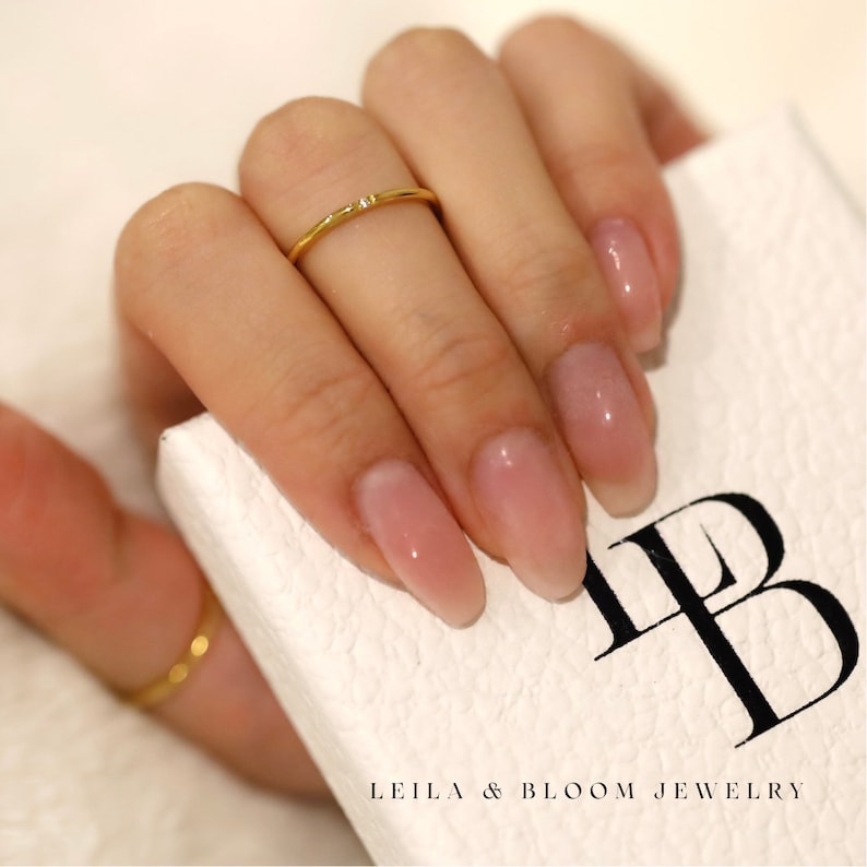 Puede incluir: Un delicado anillo de oro con un peque&ntilde;o diamante engastado en el centro. El anillo se lleva en un dedo con esmalte de u&ntilde;as rosa claro.