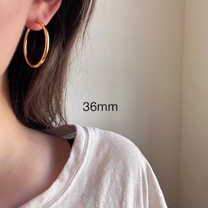 以下が含まれることがあります： 直径36mmのゴールドフープピアス。ピアスは人の耳につけられています。